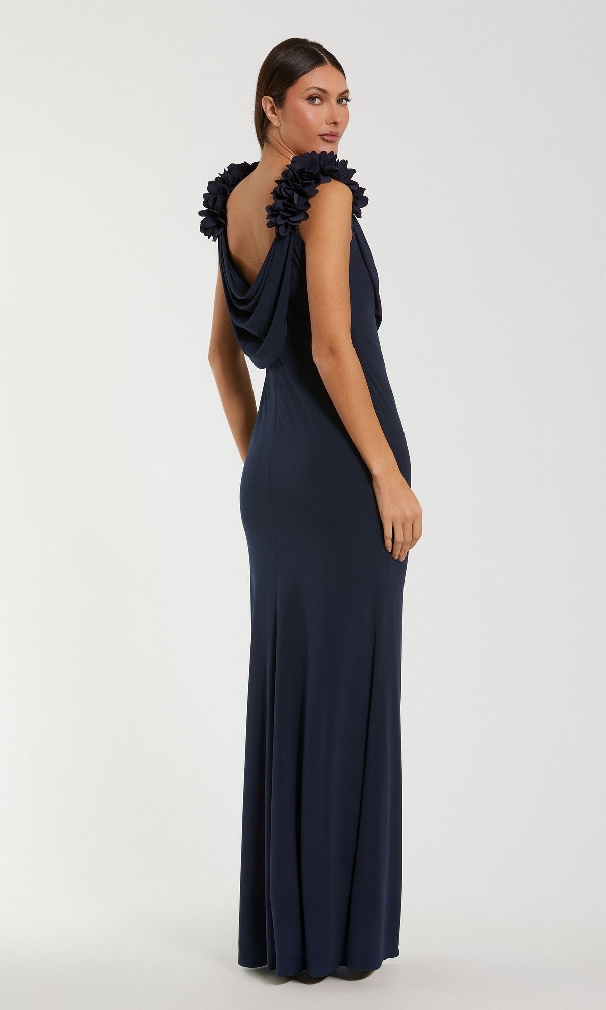 A woman models the Mac Duggal 56195 Long Formal Dress, featuring a sleeveless dark blue design with floral accents on the shoulders. She stands against a plain background, looking over her shoulder.